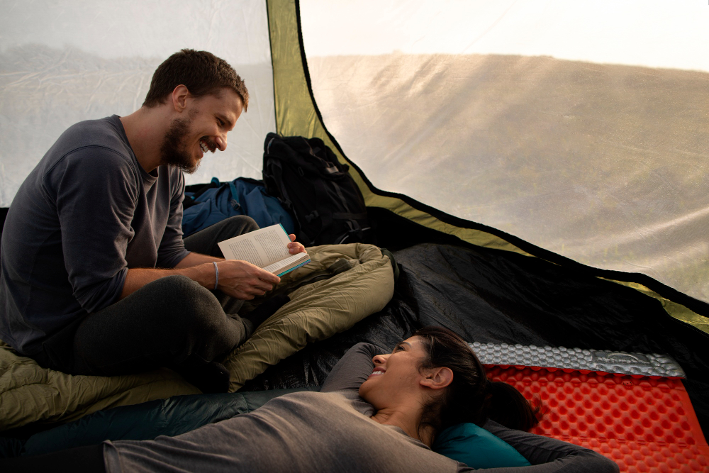 una pareja acampando utilizando sus sacos de dormir mientras el chico le lee a la chica