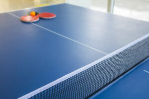 Una mesa de ping pong típica de color azul con su red, dos paletas rojas y una pelota blanca
